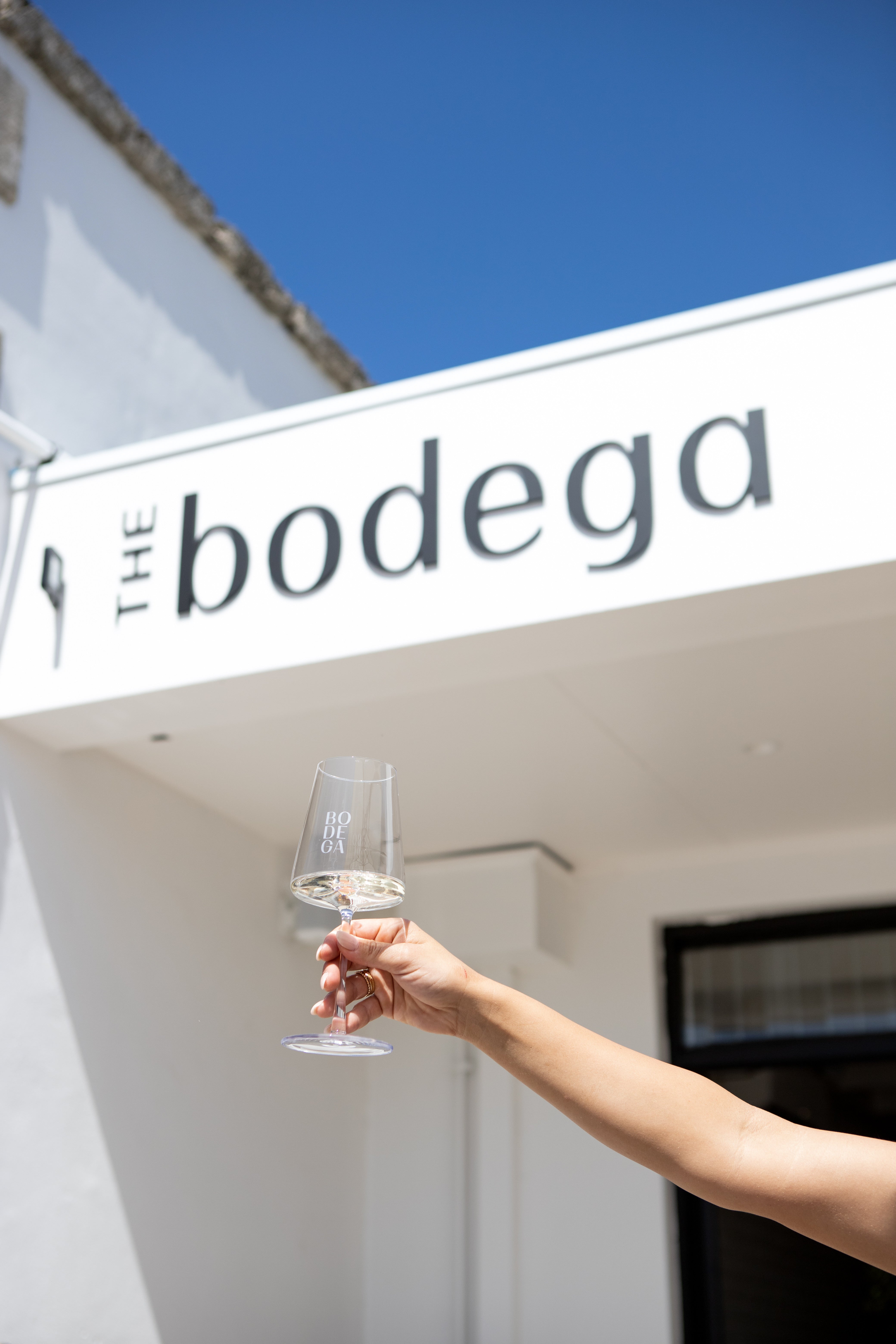 The Bodega Wine Bar and Bottle Shop sign in the sun with a bodega wine glass full of local white wine